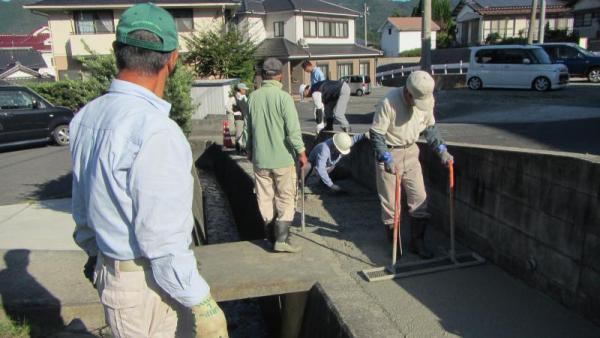 駐車場の側溝の近くの道路でコンクリートを流していく7人の男性の写真