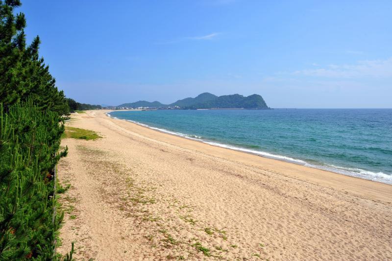 画面奥に向かって海と砂浜が続いており、その奥には山が見える写真