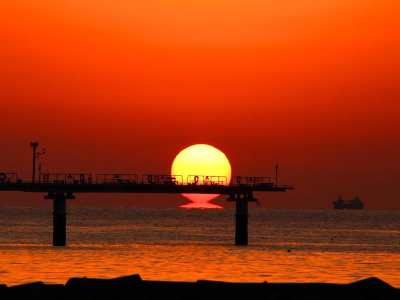 海面から突き出た橋のような建築物の向こうの水平線に太陽が沈んでいく夕暮れの写真