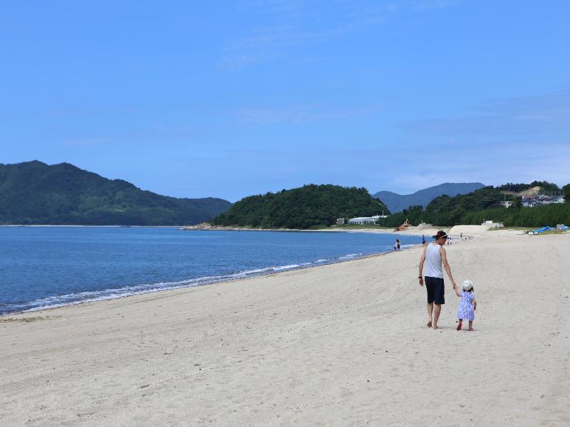 青い海とその砂浜を子供と手をつなぎ歩く人物の写真
