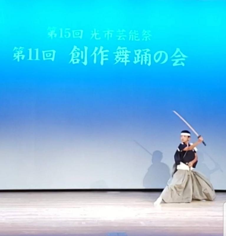 舞台上で袴を着た男性が刀を上に上げながら踊っている写真