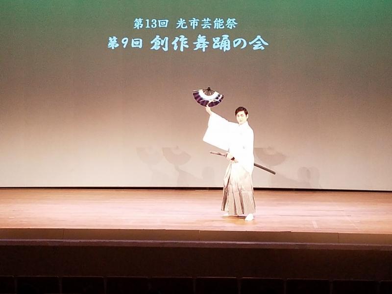 舞台上で右手に扇子、左手に刀をもっている白い袴姿の男性の写真