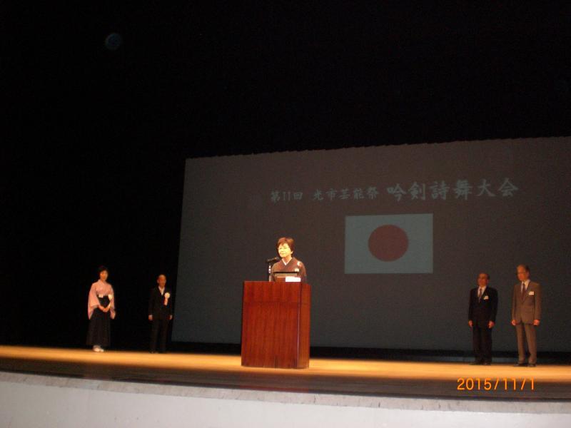 舞台上で話す着物姿の女性とそれを後ろで見ている三人の男性と一人の女性の写真