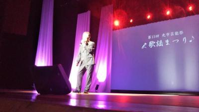 舞台上が紫のライトで照らされていて中央で男性がマイクを持って歌っている写真