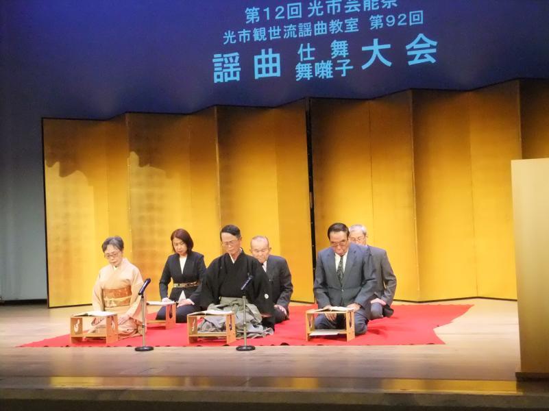 舞台上に白い文字で謡曲 仕舞 舞囃子大会と映し出されたスクリーンの前にある金屏風の前の赤い絨毯の上に正座している5人の男女の写真