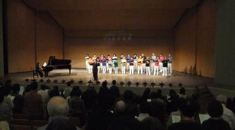白い文字で光合唱祭と映し出された舞台の端でピアノを演奏する女性とおそろいの白いズボンをはいて歌う23人の男女の前で指揮する男性の写真