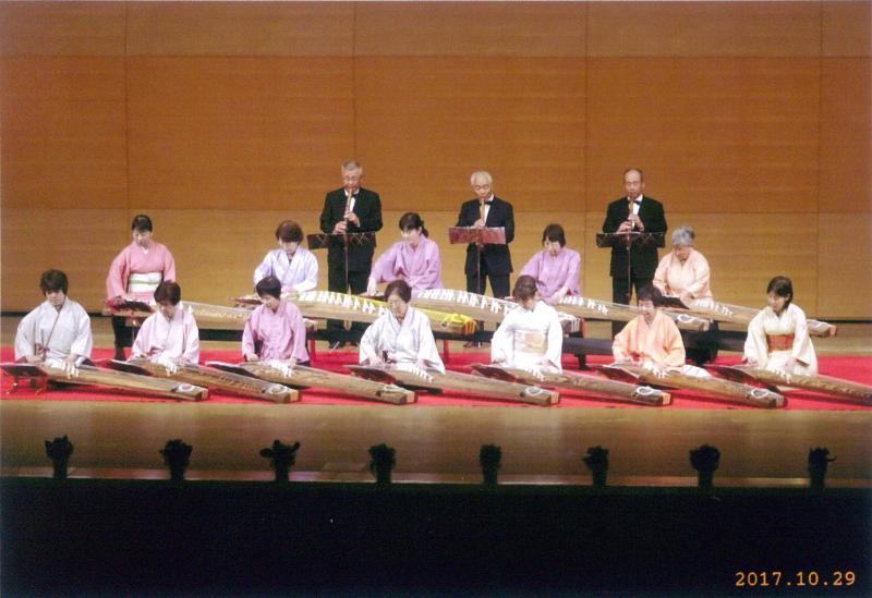 舞台上で尺八を吹く3人の男性の前で琴を演奏する12人の女性の写真