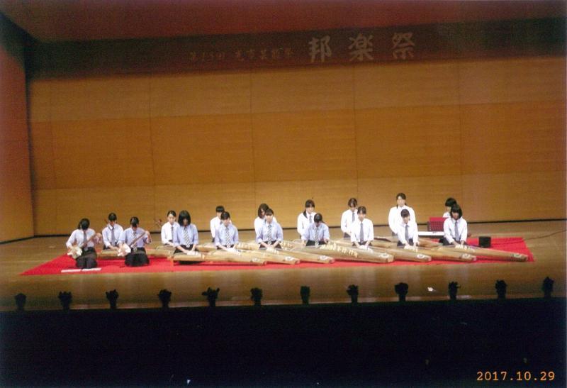 舞台の上で三味線を演奏する2人の女子学生と一緒に並んで琴を演奏する15人の女子学生の写真