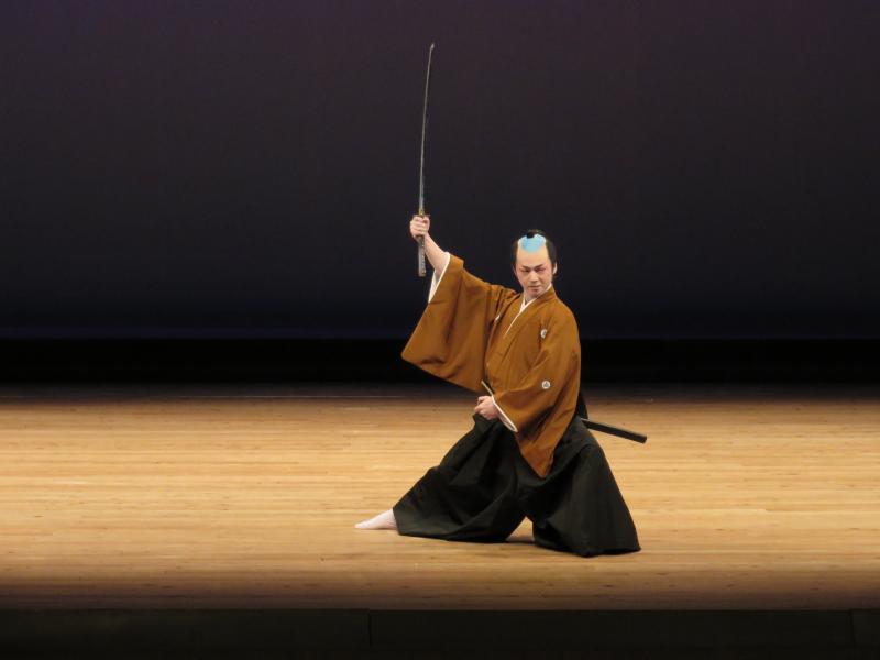 舞台の上で髷に袴姿の衣装を着て刀を掲げている男性の写真