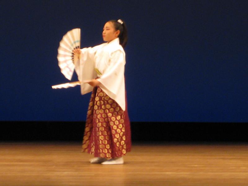舞台の上で両手に扇子を2つ持っている髪を一つに結わった白い袴姿の女の子の写真