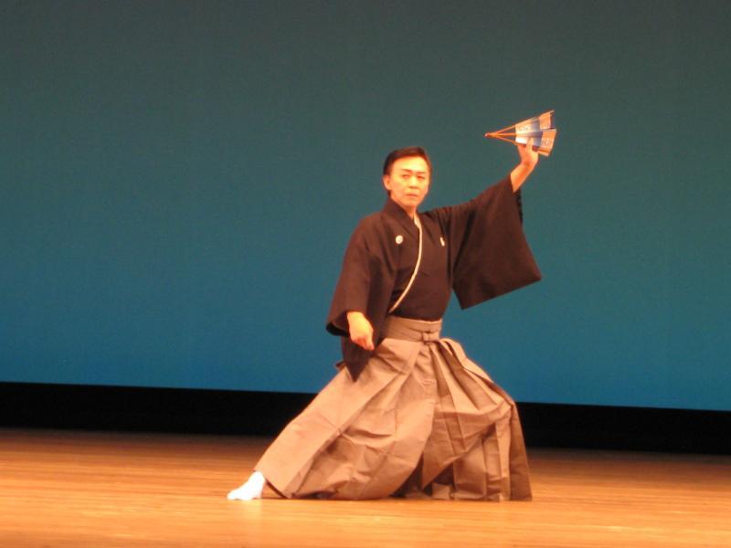 舞台の上で扇子を左手に持って顔より上の位置に挙げている黒い袴姿の男性の写真