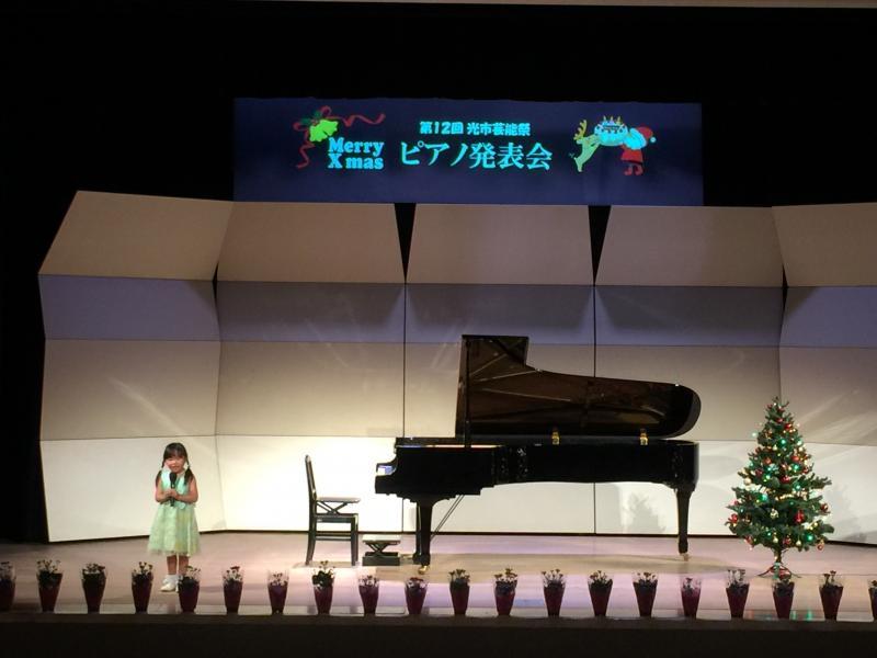 舞台上の第12回光市芸能祭ピアノ発表会と映し出されたスクリーンの前に置いているピアノの横に立ってマイクを持っている黄緑色のワンピースを着た女の子の写真