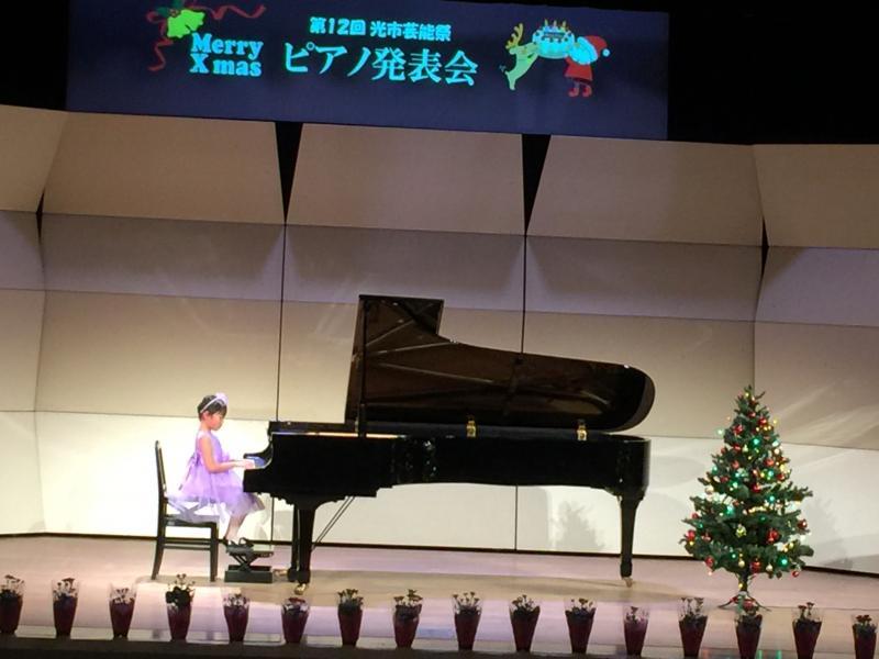 舞台上の第12回光市芸能祭ピアノ発表会と映し出されたスクリーンの前に置いているピアノに座って演奏している薄紫色のドレスを着た女の子の写真