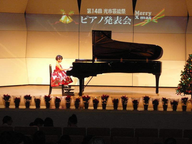 端に等間隔に鉢植えが置かれている舞台の真ん中でピアノを演奏する赤い花柄のドレスを着た女の子の写真
