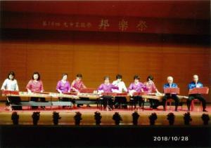 舞台の上で琴を演奏するそれぞれピンク・赤・白の衣装を着た8人の女性と尺八を演奏する青い服の2人の男性の写真