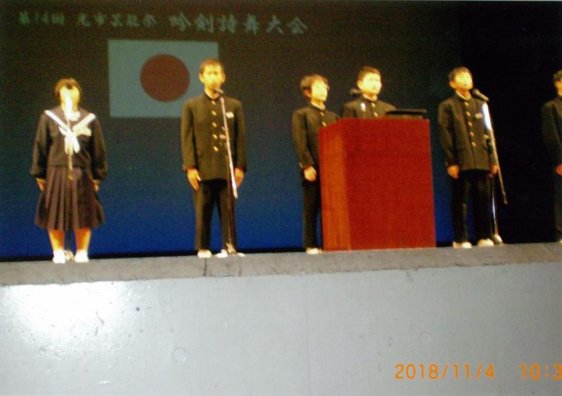 吟剣詩舞大会という文字と日本国旗が映し出されたスクリーンを背景に舞台上で立っている制服姿の6人の学生の写真