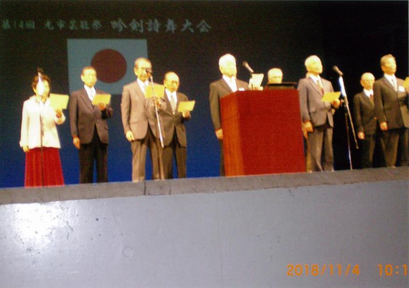吟剣詩舞大会という文字と日本国旗が映し出されたスクリーンを背景に舞台上で黄色い紙を持って立っている9人の男女の写真