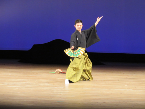 舞台の上で広げた扇子を持って舞を舞っている袴姿の一人の男性の写真