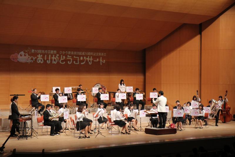 白い背広を着た男性の指揮にあわせて楽器を演奏する28名の男女の写真