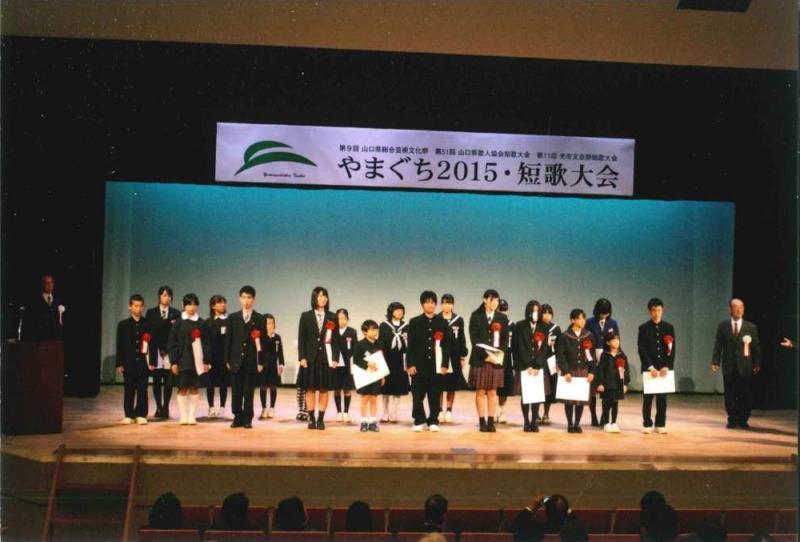 舞台上に「やまぐち2015・短歌大会」と書かれた看板の下で賞状を持って立っている20人の男女の学生の写真