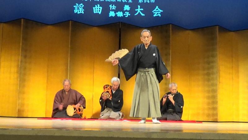 舞台の上で鼓を打つ三人の男性の前で扇子を持って舞を踊る袴姿の男性の写真