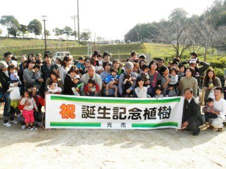 「祝誕生記念植樹」と書かれた幕の後ろに大勢の参加者たちが並んでいる集合写真