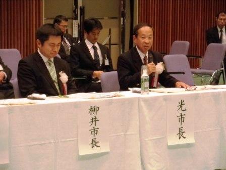 光市長と柳井市長が並んで座っていて、光市長がマイクを持って話をしている写真