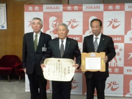 田村 敏雄さんが表彰状を持って中央に立っていて、その横に市長と関係者1名が並んでいる写真