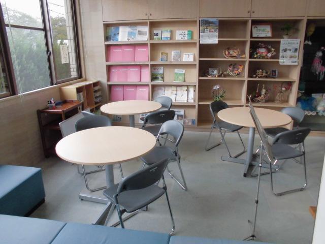 奥の壁側に本やパンフレットが並ぶ本棚と、丸い机と椅子がそれぞれ3脚設置されている交流スペース室内の写真