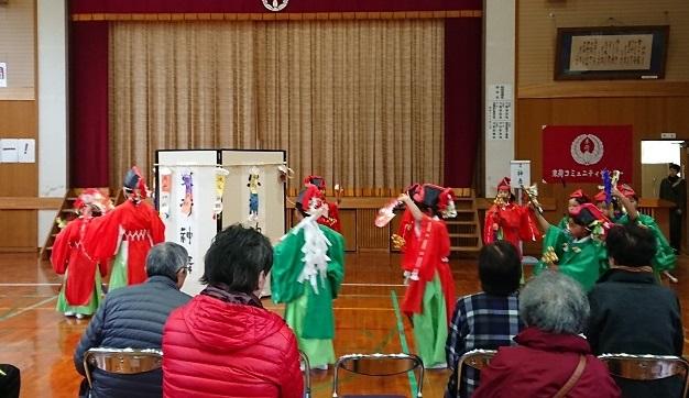 体育館で束荷小学校の生徒が赤や緑の衣装を着て真ん中に置いてある屏風の周りを踊っている様子を観客が見ているアップの写真