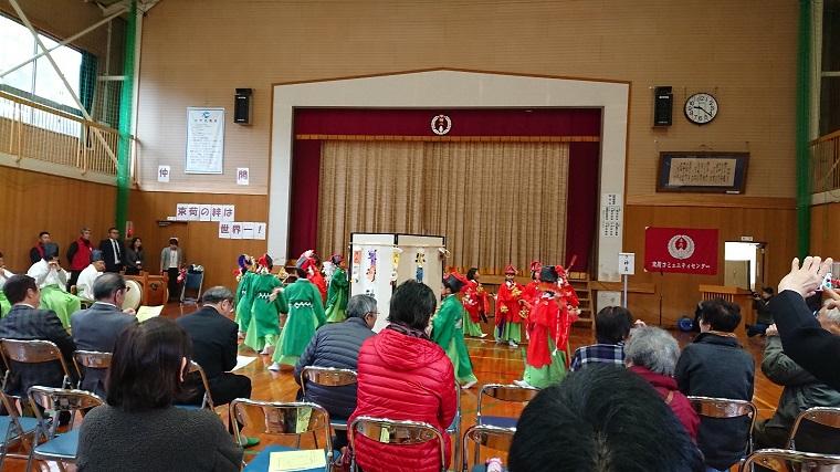 体育館で束荷小学校の生徒が赤や緑の衣装を着て真ん中に置いてある屏風の周りを踊っている様子を観客が見ている写真