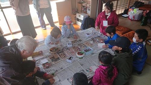新聞紙が敷かれた机の上で手作りのハンコなどを使って年賀状を作成している子供たちの写真