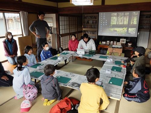 和室の机の上に新聞紙を引き青版と消しゴムを置いて子供たちが指導者の話を聞いている写真