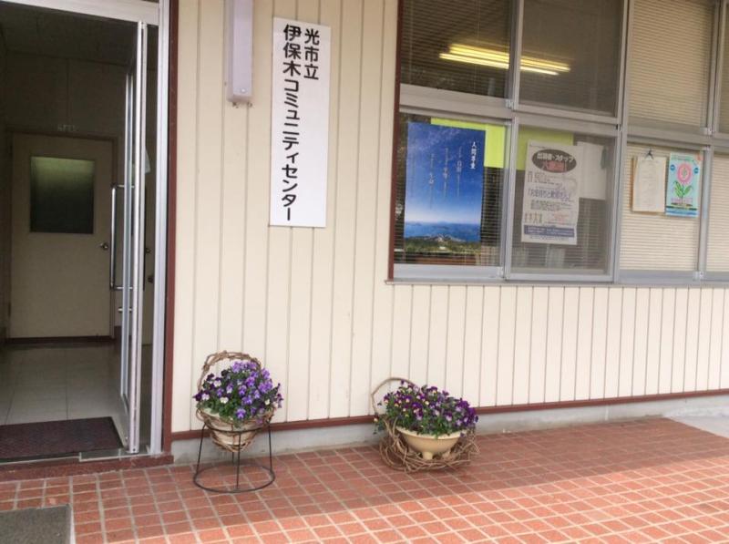 外側の入口横の壁に光市立伊保木コミュニティセンターと書かれた表札があり、その下に花が植えられた鉢が2つ置かれてある写真