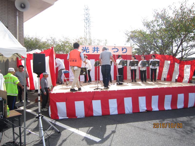 紅白の幕が張り巡らされた舞台上で、各々の譜面台の前に女性4人が立っており、後ろ姿の男性とオレンジ色のベストを着た男性が写っている写真