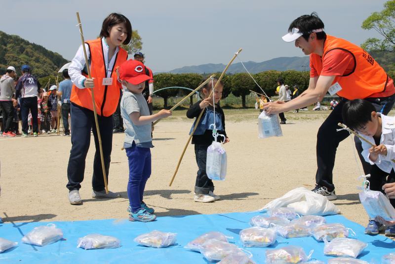 運動場にて小さな子供たちが手に手作りの竿でブルーシートの上に置かれた景品の入った袋を吊り上げ、男性隊員に渡している写真