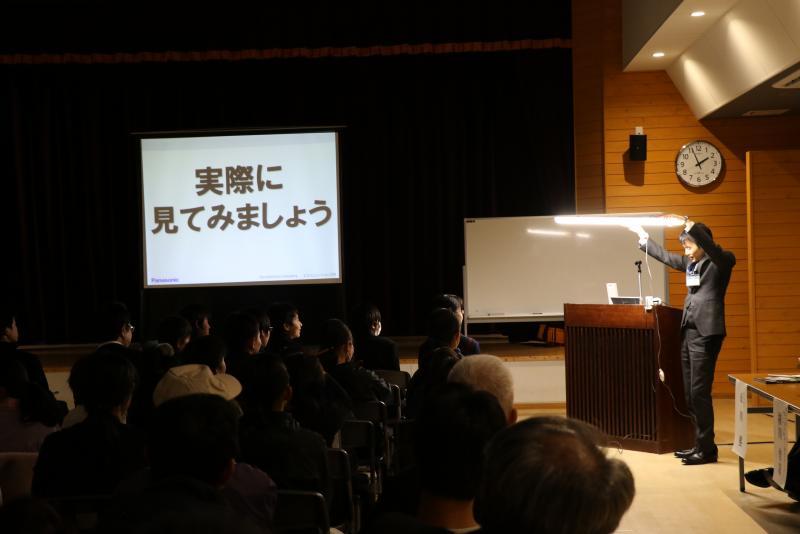スクリーンに「実際に見てみましょう」と映し出され、聴衆の前で田村技師が説明している写真