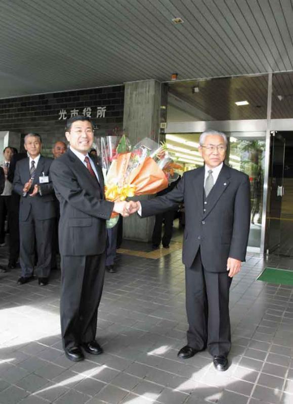 花束を持つ初代市長の末岡 泰義さんと握手をする男性の写真