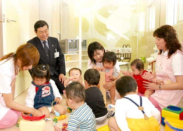梅田病院（虹ヶ浜）内の病後児保育所「Loving HUG」でたくさんの子供を保育士3人で預かっている様子を背広姿の男性が見ている写真