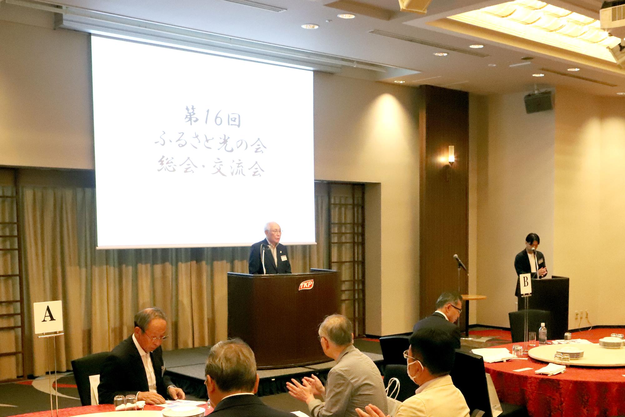 円卓が並ぶ室内で、前に立って話す男性と清聴する人たちの写真