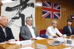 4人の人物が横に並んで座っており、その後ろに光と書かれた幕と英国国旗が飾られている写真