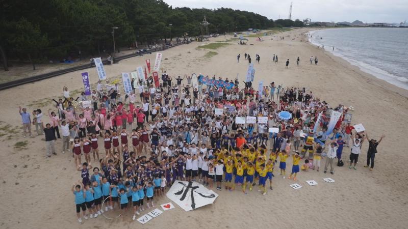 砂浜で撮影に参加した市民たちが一か所に集まりその様子を上空から撮影した写真
