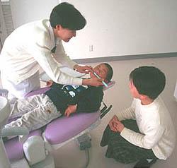 歯医者のリクライニングする椅子に横になっている男児の歯を磨き保護者に歯磨き指導を行う歯科医の写真