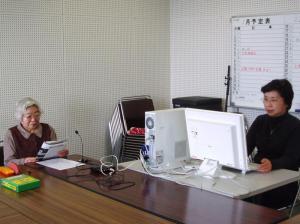 予定表の書いているホワイトボードがある部屋の中で白いパソコンで作業するメガネの女性と長机の前で冊子を読んでいる白髪の女性の写真