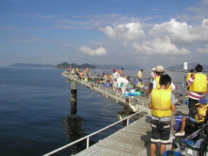 遠くに島が見える海の上のくの字型の桟橋の上で黄色いライフジャケットを着用して釣りをしている大勢の人々の写真