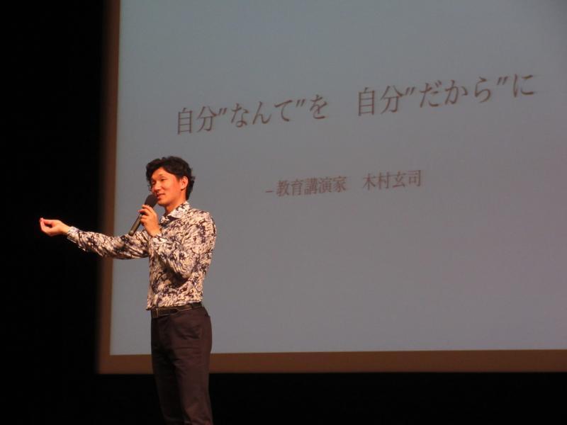 柄モノのシャツを着た男性がマイクを片手にステージ上で話している写真