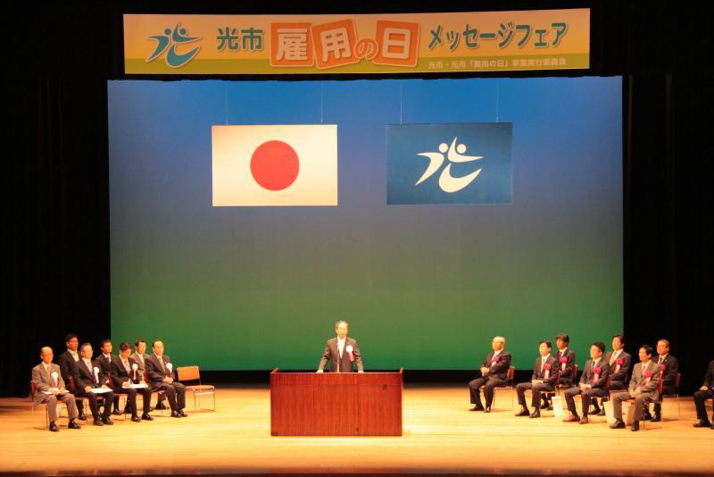 国旗と市町村旗が掲げられたステージに立って話している男性の写真