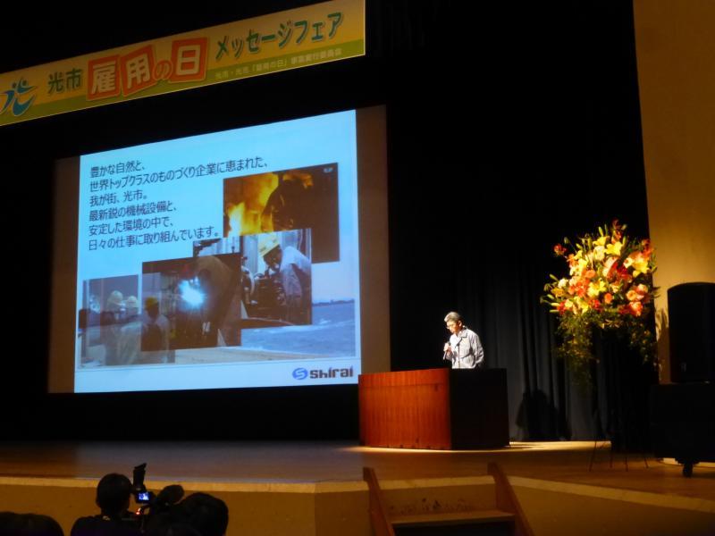 ステージ上のスクリーンに白井興業株式会社のプレゼンのスライドが映し出されている写真